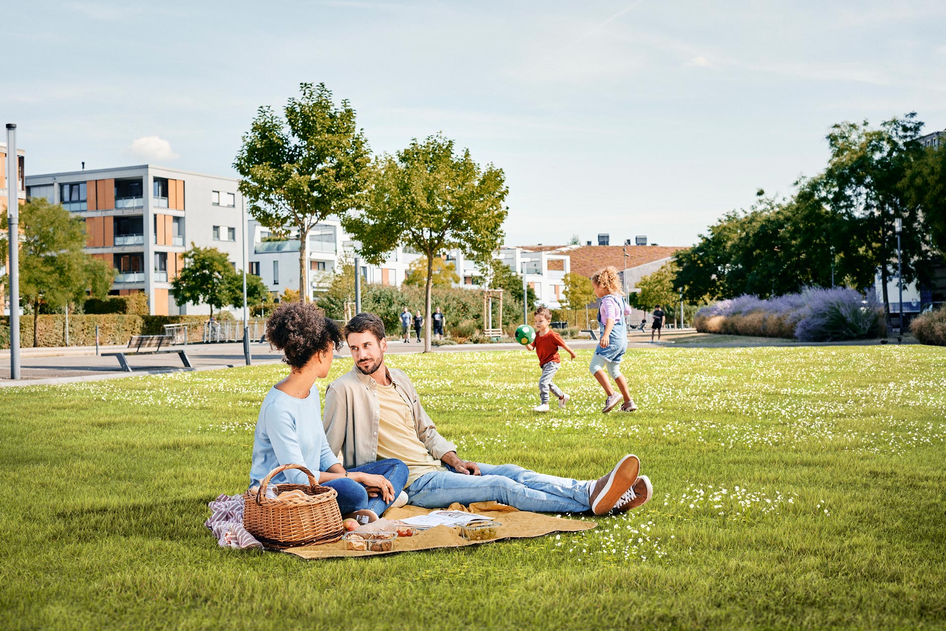 Ein junges Paar sitzt auf einer Decke auf einer grünen Parkwiese. Im Hintergrund spielen zwei Kinder Ball.