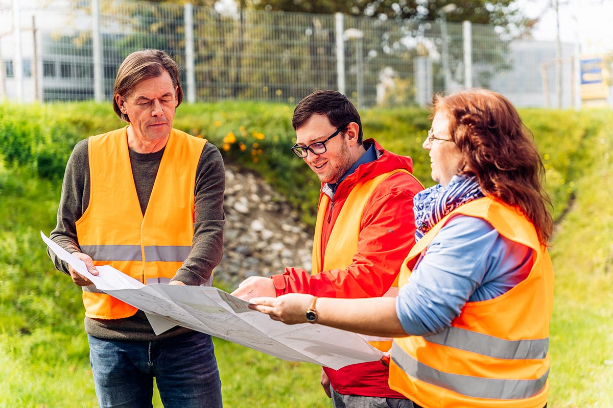 Drei Projektbeteiligte vom Bau einer Versickerungsmulde schauen in orangefarbenen Warnwesten auf die Bauskizze. 