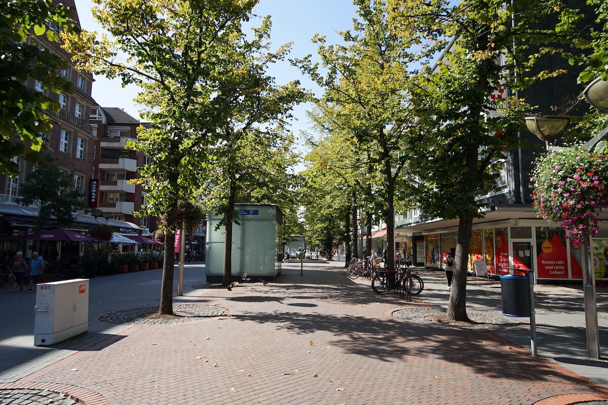 Zu sehen ist eine Einkaufsstraße, viele Einzelhandelgeschäfte und ein breiter Fußgängerweg welcher sich mit Bäumen schmückt.