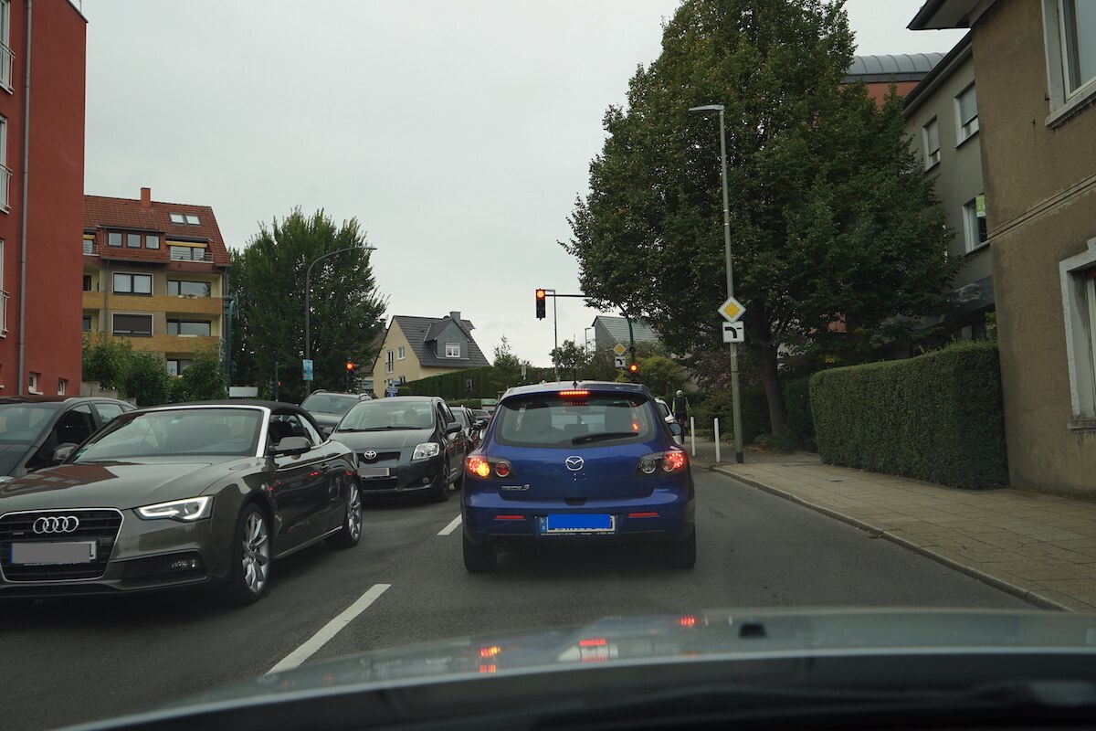 Zu sehen sind mehrere Autos die vor einer roten Ampel stehen. Im Fokus ein blaues Auto. 