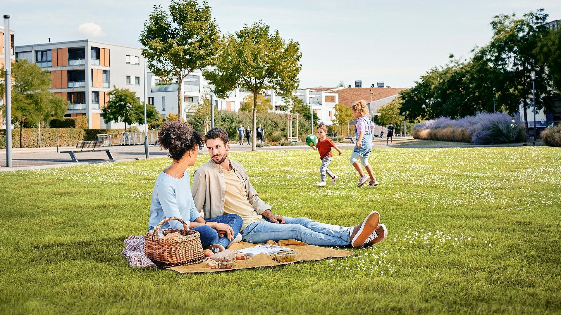 Ein junges Paar sitzt auf einer Decke auf einer grünen Parkwiese. Im Hintergrund spielen zwei Kinder Ball.